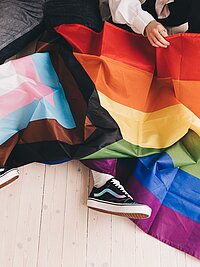 Zu sehen ist eine Regenbogenflagge die über den Beinen von zwei Menschen ausgebreitet liegt.