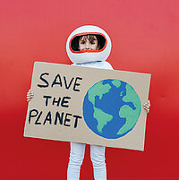 Ein Mädchen in einem Astronautenkostüm mit Helm steht vor rotem Hintergrund und hält ein selbsgebasteltes Schild in den Händen mit einer Weltkugel und dem Text "Save the Planet"