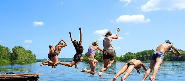 7 Jugendliche springen vermutlich von einem Steg in einen See, man sieht sie von hinten